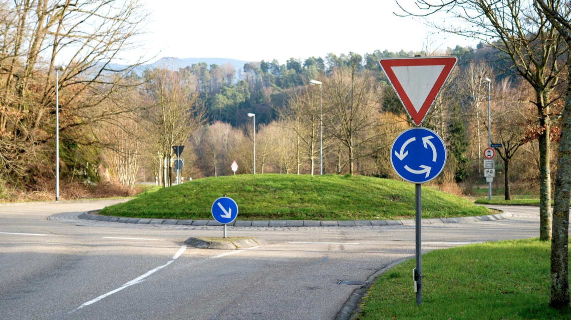 Ein Kreisverkehr mit begrüntem Mittelbereich. Im Vordergrund stehen ein dreieckiges Vorrang-geben-Schild sowie ein blaues Kreisverkehrszeichen mit Pfeilen zur Fahrtrichtung. Weitere Verkehrszeichen befinden sich entlang der Straße