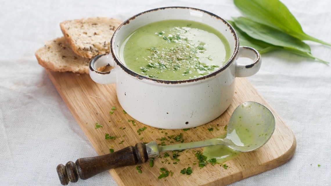 Auf einem Holzbrett auf einem Tischtuch steht ein Holzbrett, auf dem ein weißer, kleiner Suppenteller mit Bärlauchsuppe steht. Daneben liegen zwei Scheiben Brot, ein Löffel und Bärlauchblätter