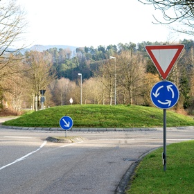 Ein Kreisverkehr mit begrüntem Mittelbereich. Im Vordergrund stehen ein dreieckiges Vorrang-geben-Schild sowie ein blaues Kreisverkehrszeichen mit Pfeilen zur Fahrtrichtung. Weitere Verkehrszeichen befinden sich entlang der Straße