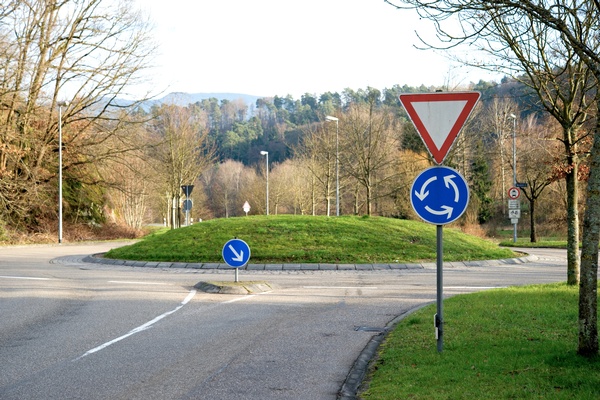 Ein Kreisverkehr mit begrüntem Mittelbereich. Im Vordergrund stehen ein dreieckiges Vorrang-geben-Schild sowie ein blaues Kreisverkehrszeichen mit Pfeilen zur Fahrtrichtung. Weitere Verkehrszeichen befinden sich entlang der Straße