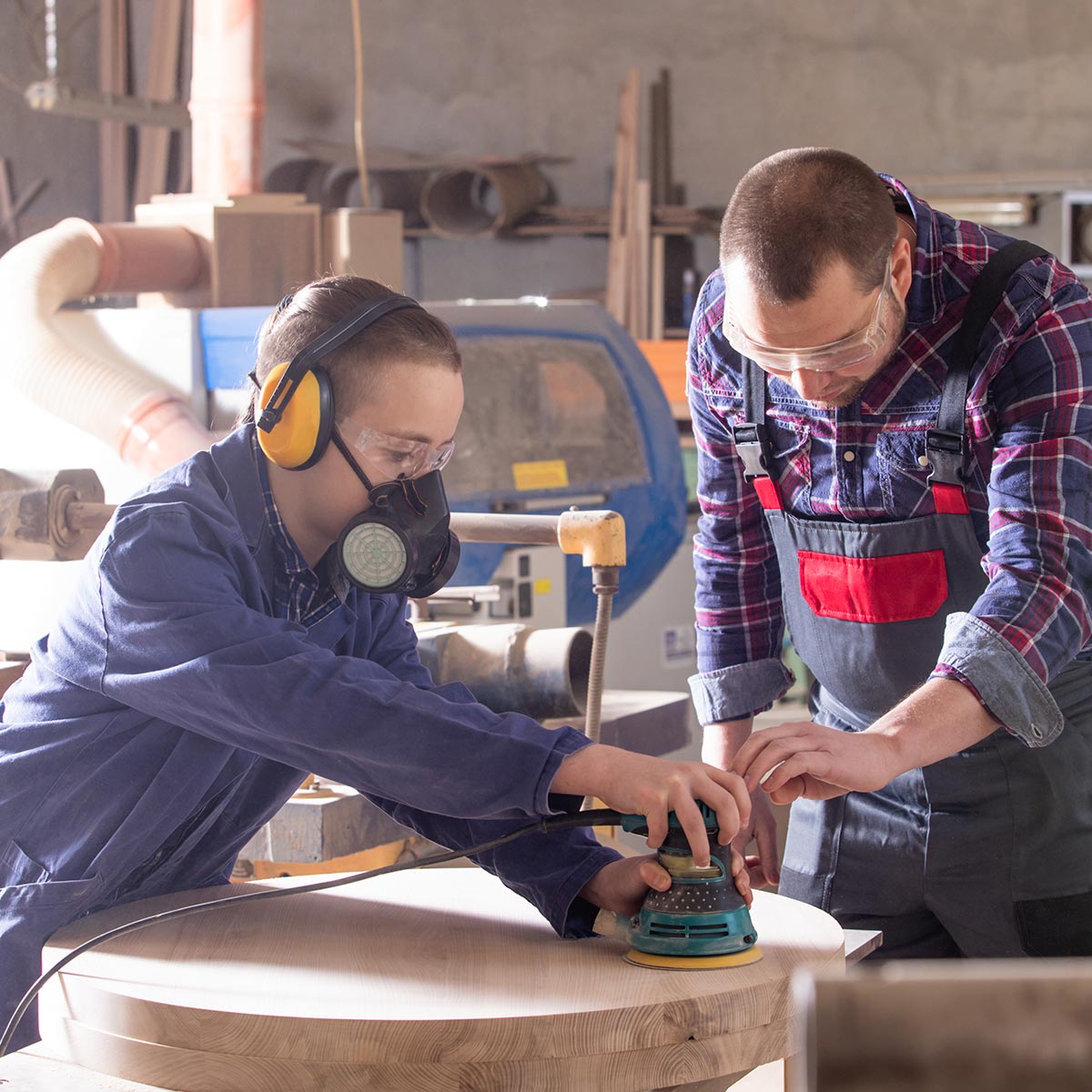 Fachkraft begleitet Lehrling mit Schutzausrüstung beim Schleifen eines Holzstücks in der Werkstatt.
