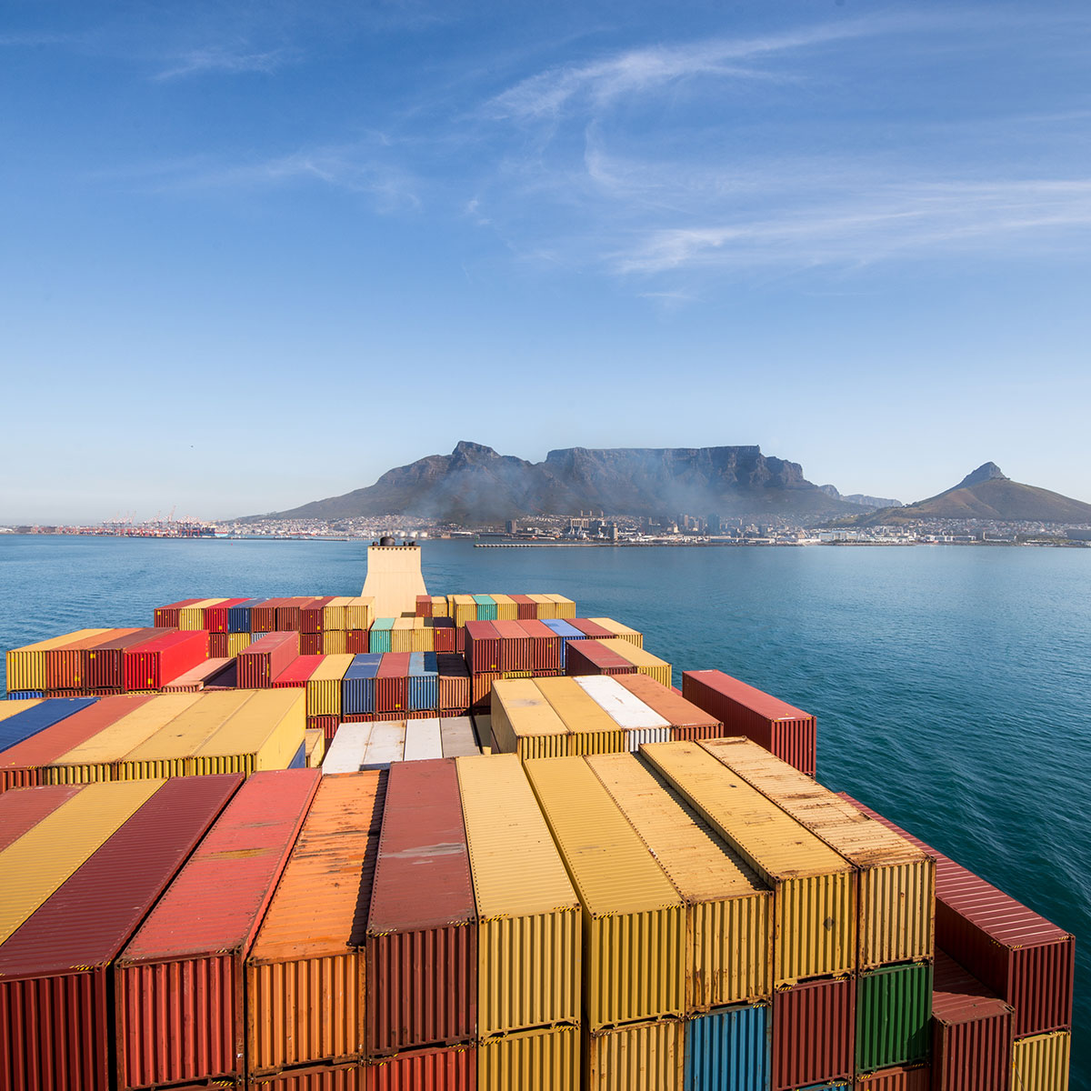 Blick von einem Containerschiff auf bunte Schiffscontainer im Vordergrund, dahinter das Meer und den Tafelberg in Kapstadt, Südafrika, bei klarem, blauem Himmel.