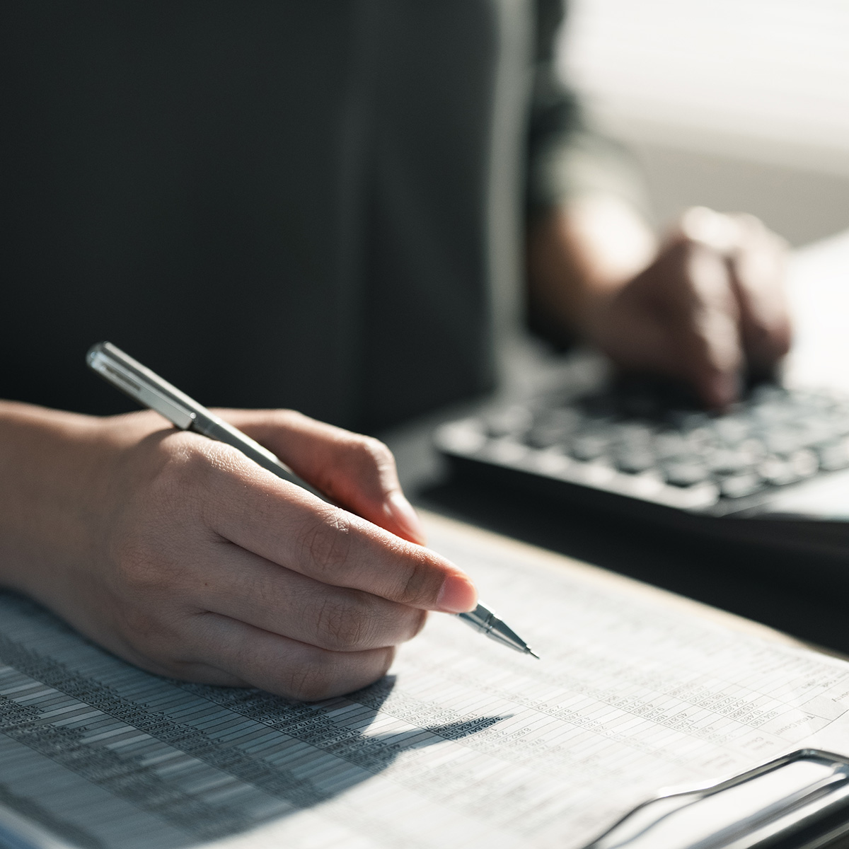 Person prüft Zahlen auf einer Liste und nutzt einen Taschenrechner am Schreibtisch.