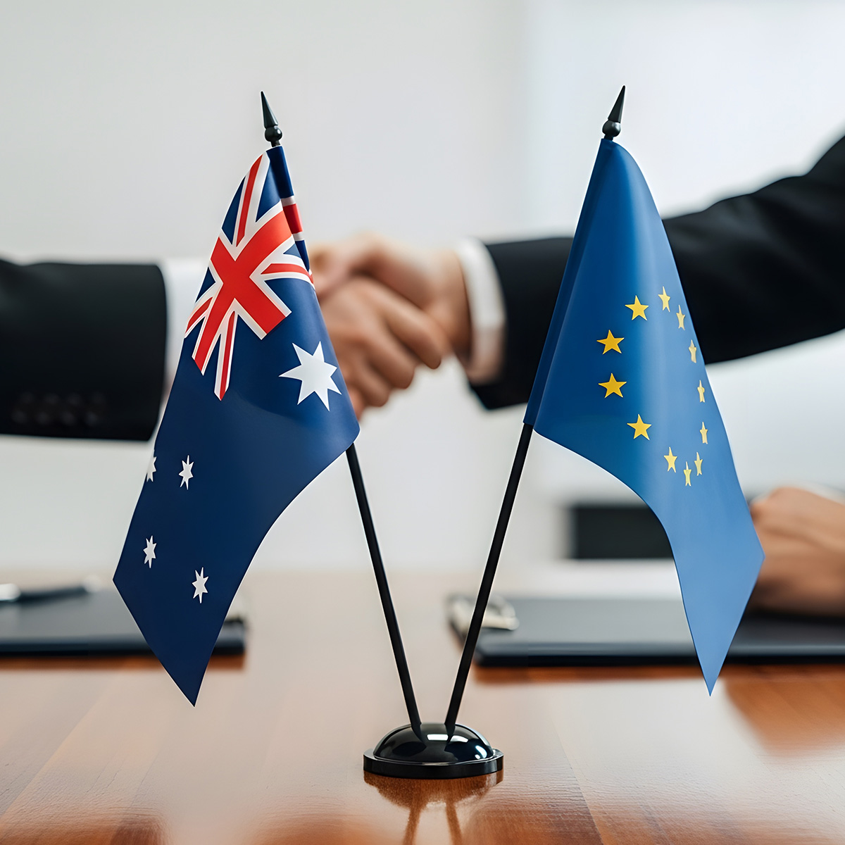Australien- und EU-Flaggen auf Konferenztisch bei Geschäftstreffen mit Handschlag im Hintergrund.