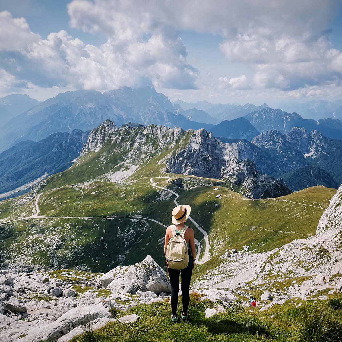 Person mit Rucksack blickt von einem Bergweg auf eine weitläufige Alpenlandschaft