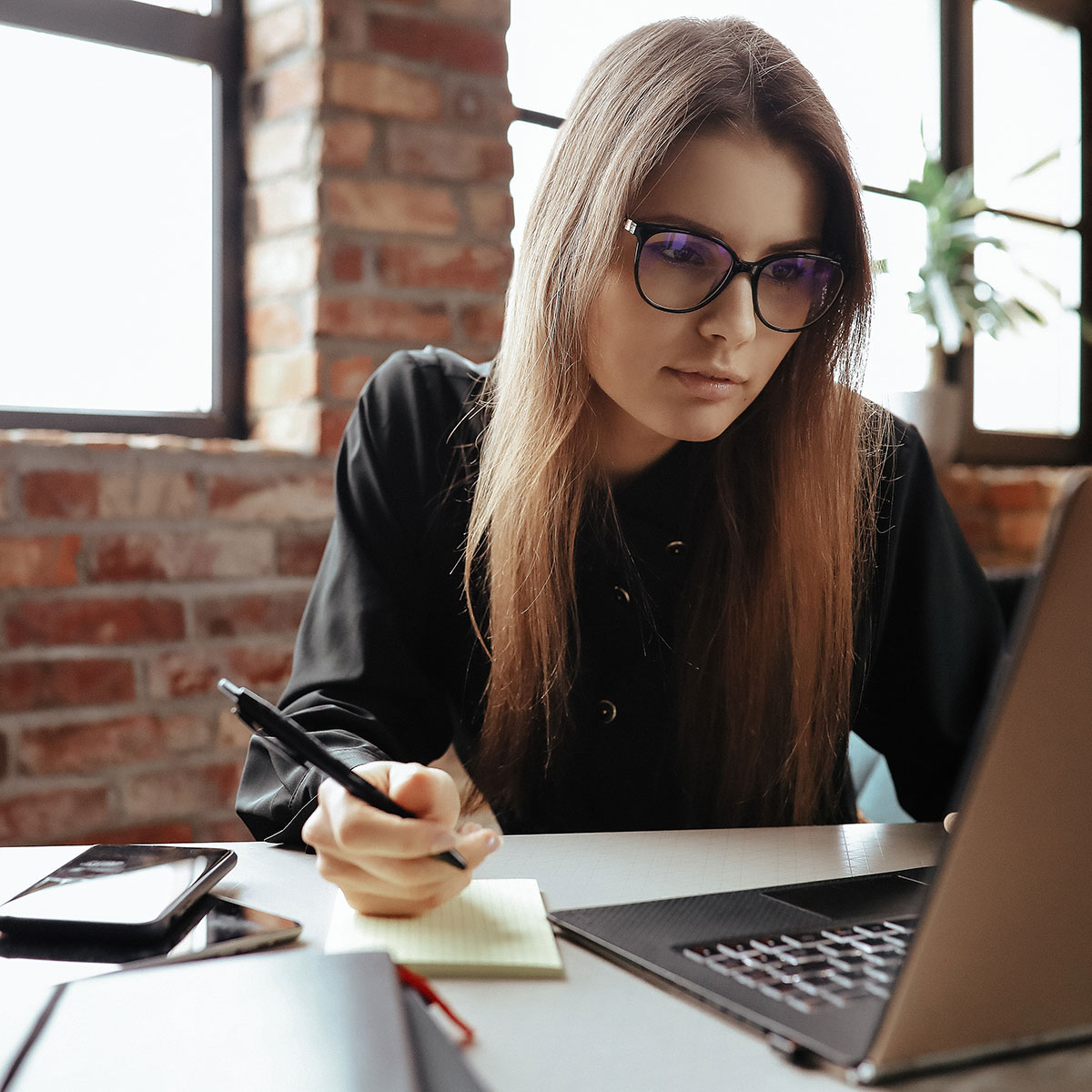 Frau mit Brille arbeitet im modernen Büro am Laptop und macht Notizen am Schreibtisch.